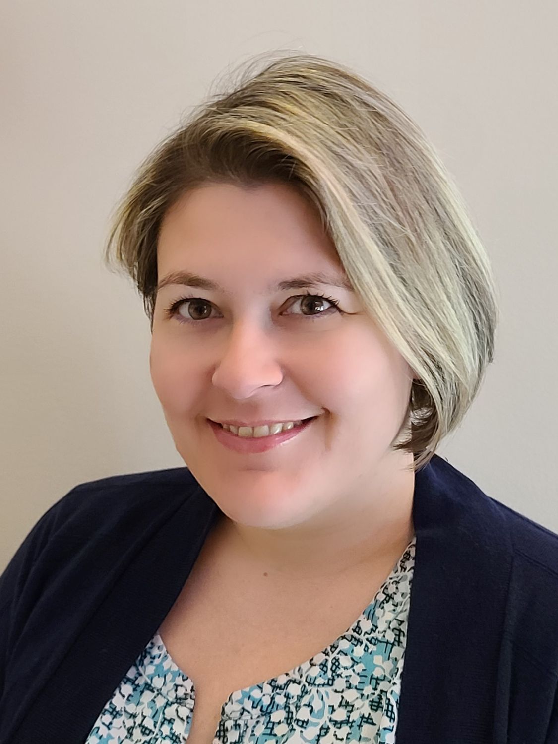 Woman with short blonde hair smiling, wearing a blue sweater and floral shirt.