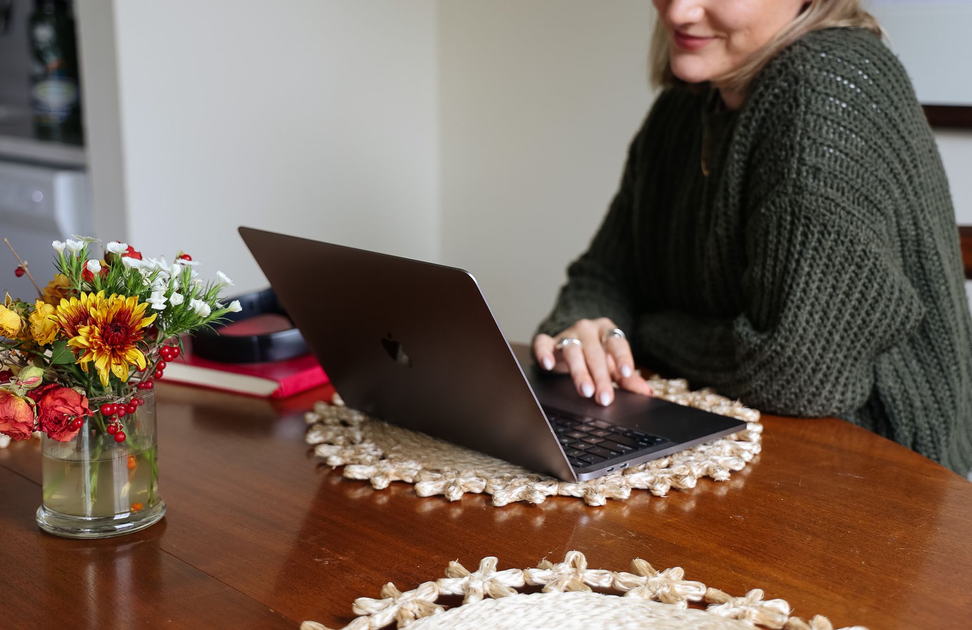 Woman using laptop at a table, wearing a green sweater. Flowers and a placemat sit nearby.