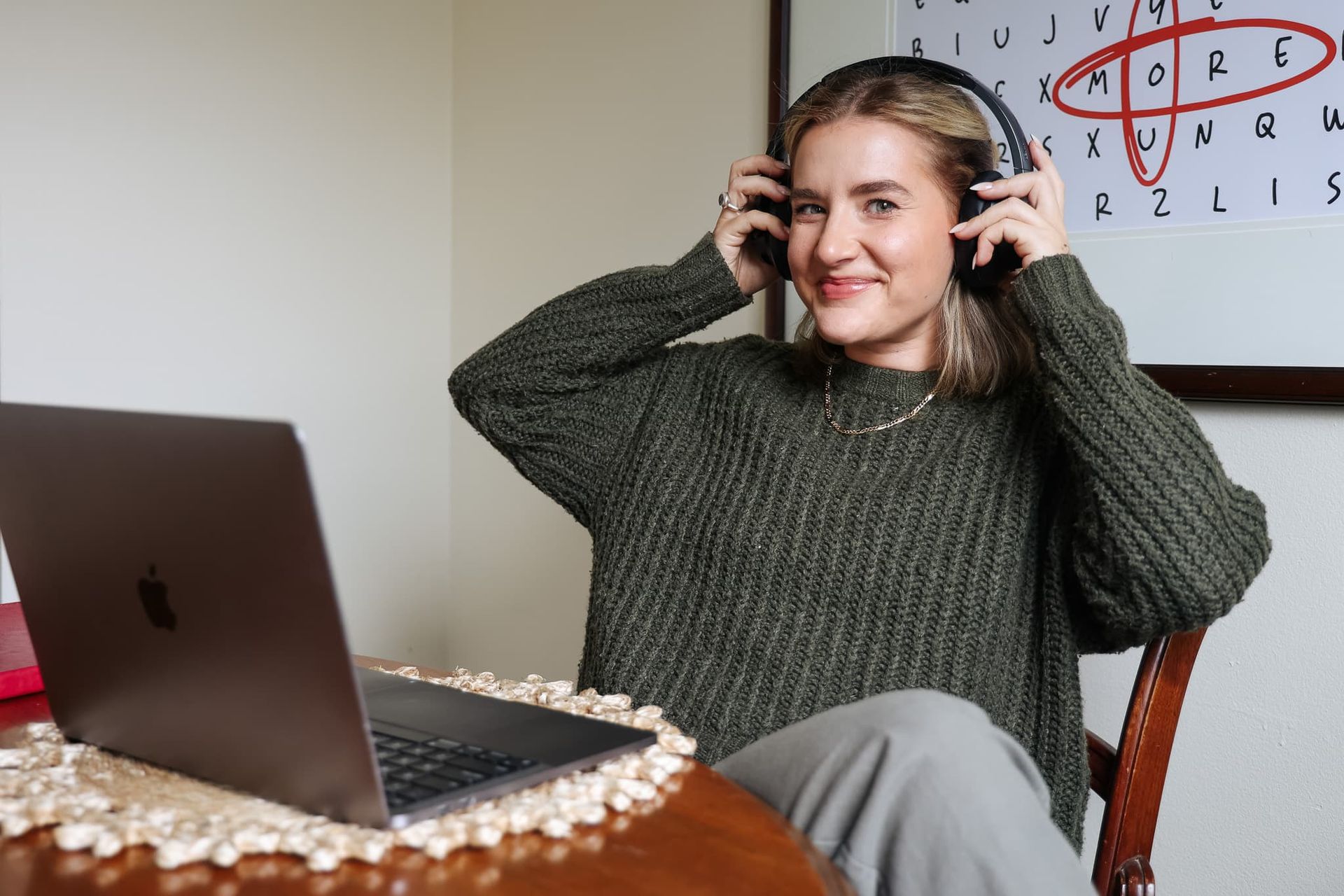 Woman wearing headphones at a table, smiling beside an open laptop in a home office
