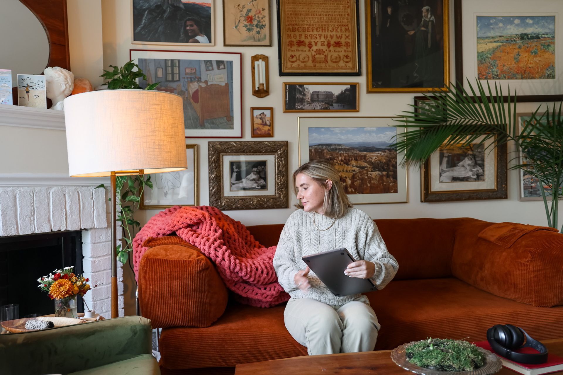 Woman sits on orange couch, holding a laptop in a cozy, art-filled living room.