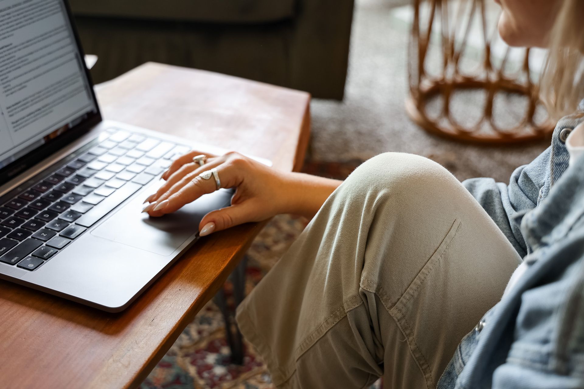 Woman using laptop, hands on trackpad. Wooden table, neutral-toned pants, denim jacket.