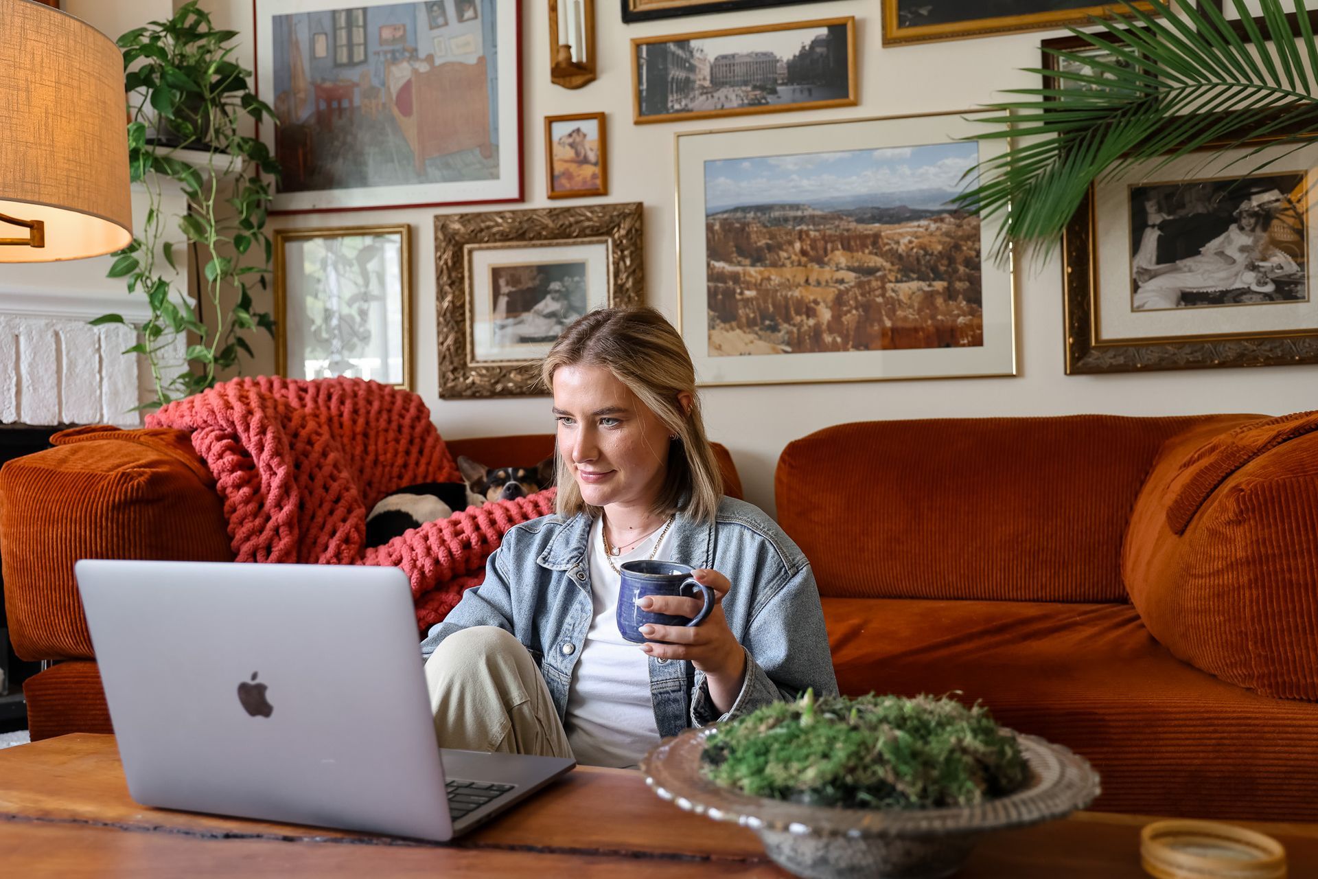 A person sits on an orange couch holding a mug while looking at a laptop, surrounded by framed art on a wall.