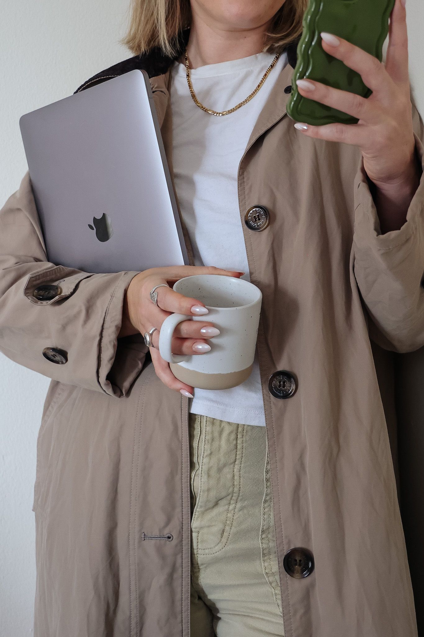 A person in a beige trench coat holds a laptop, a mug, and a green smartphone, wearing a white shirt and gold necklace.
