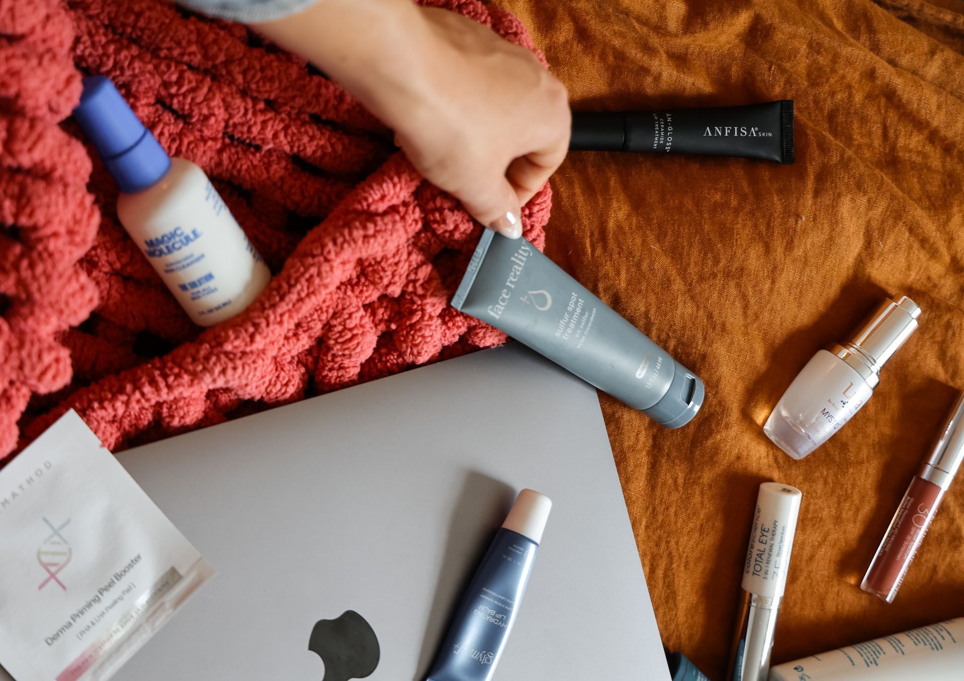 Person's hand reaching for gray face cream on a blanket with beauty products and a laptop.