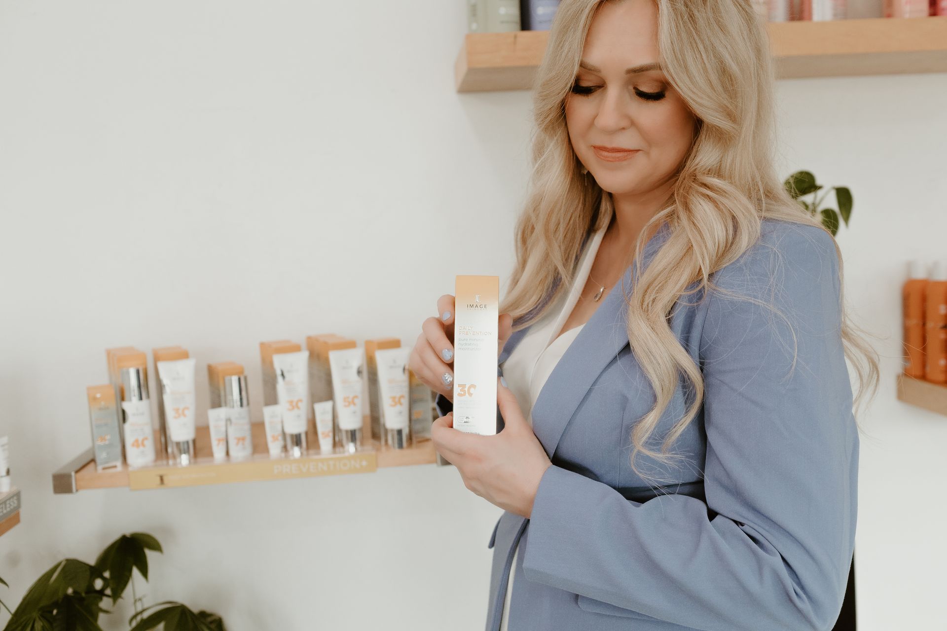 Woman in a blue blazer examines products, seated near a window and mirror with floral accents.