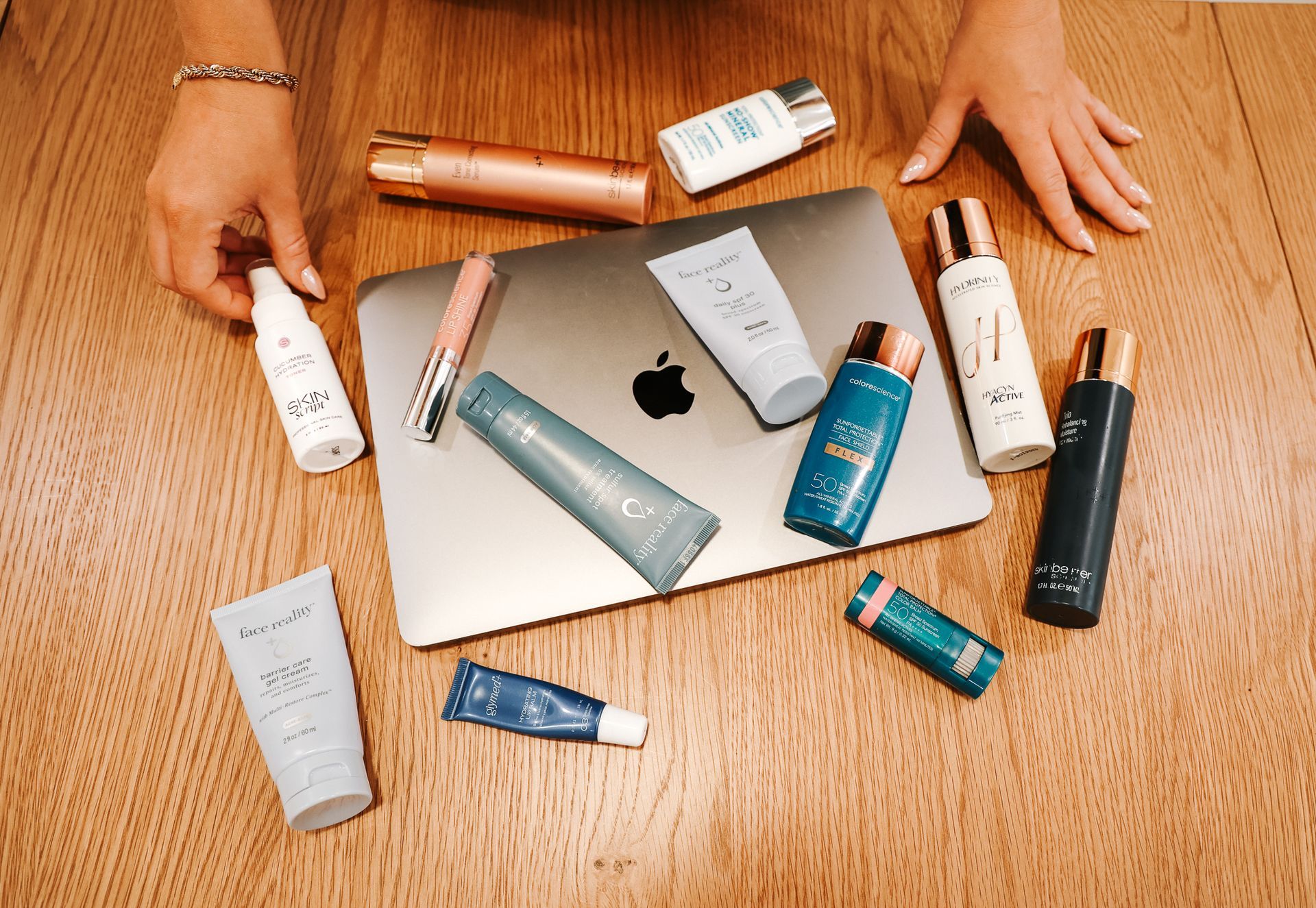 Hands surrounding a laptop with scattered skincare products on a wooden floor
