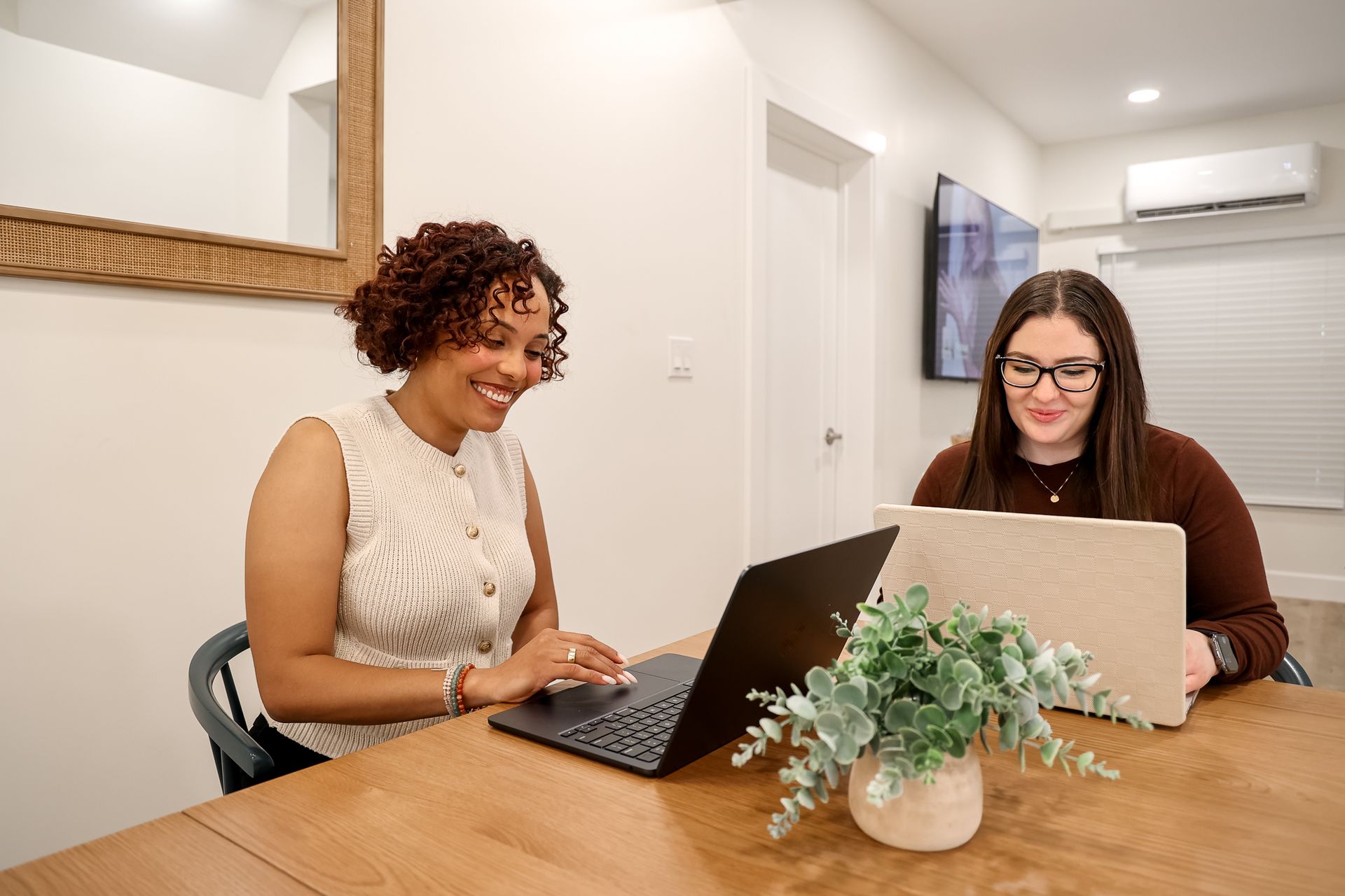Two women working on laptops at a wooden table in a well-lit room; one smiles, the other looks focused.