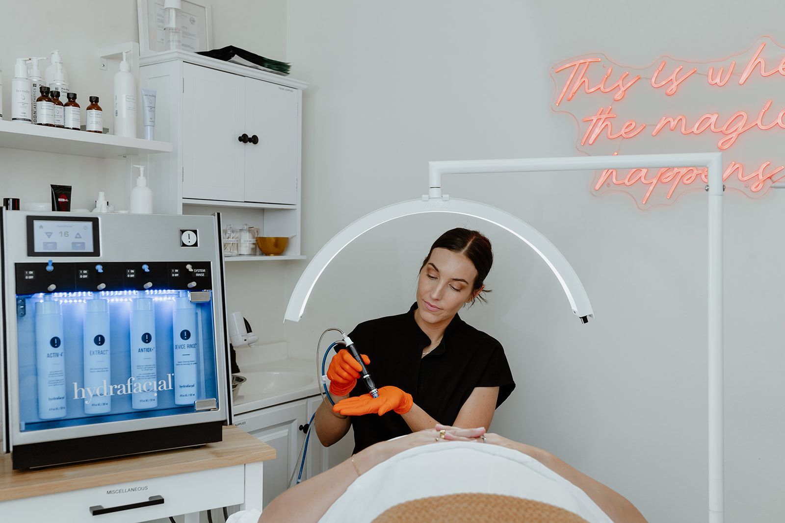 Person performing a facial treatment in a bright spa room with a glowing ring light and skincare machine