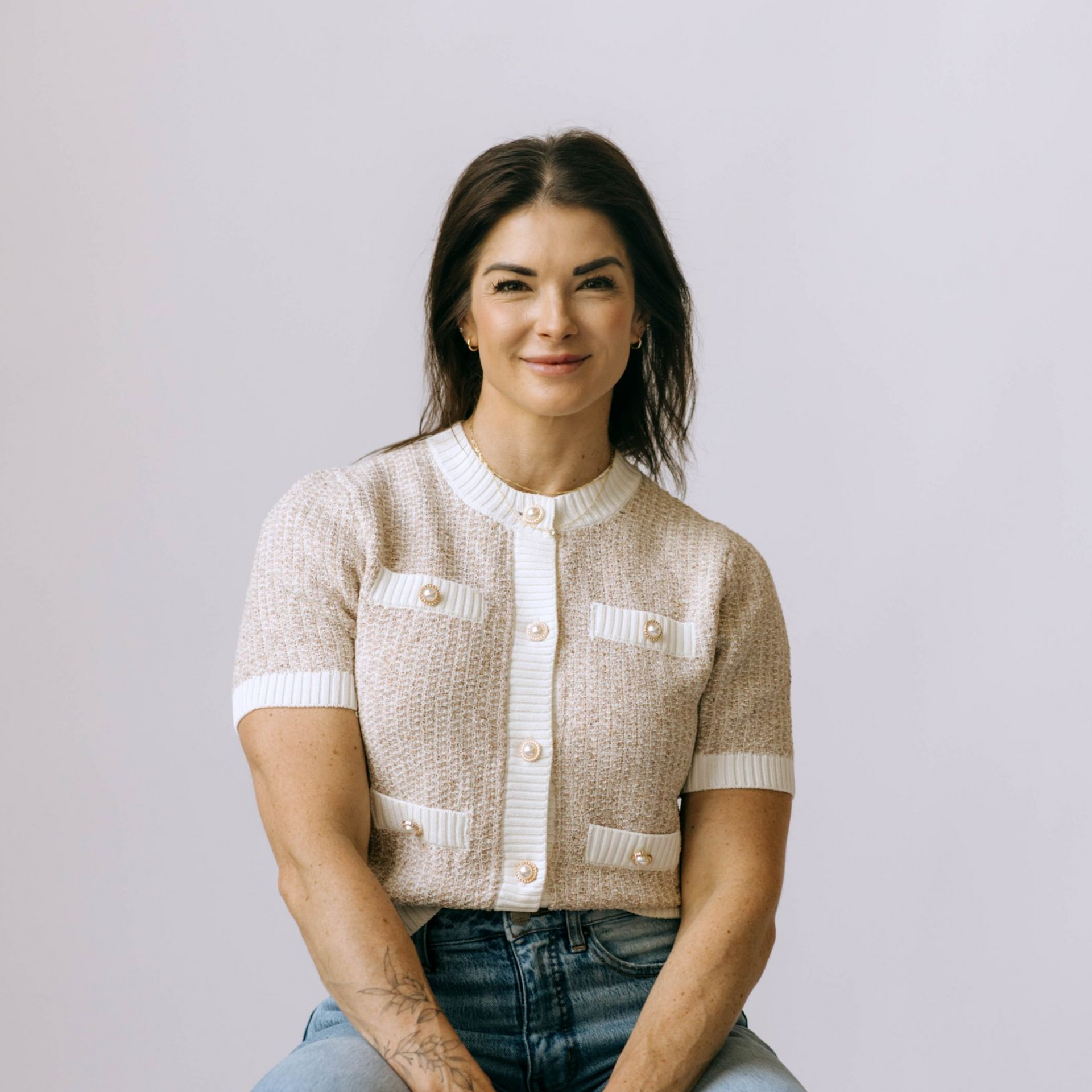 Woman wearing a cream cardigan and jeans, smiling, seated against a white backdrop.