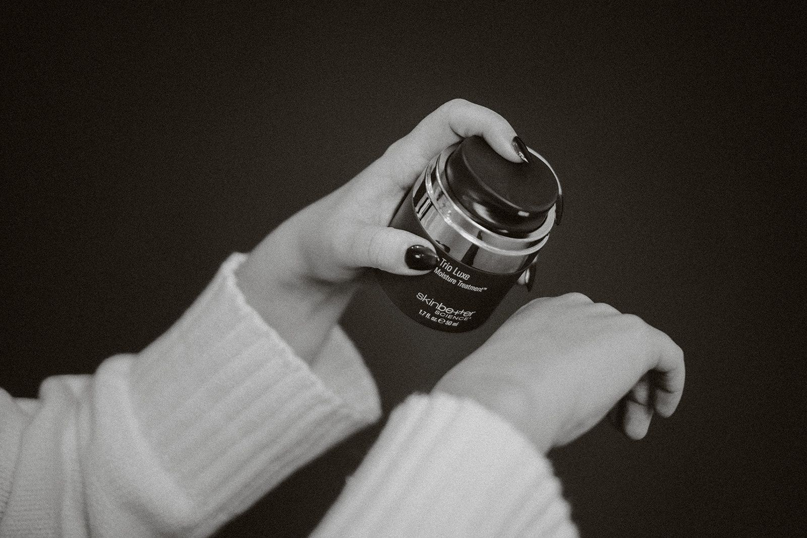 A person's hands holding a jar of cream, applying it to the wrist. Black and white.