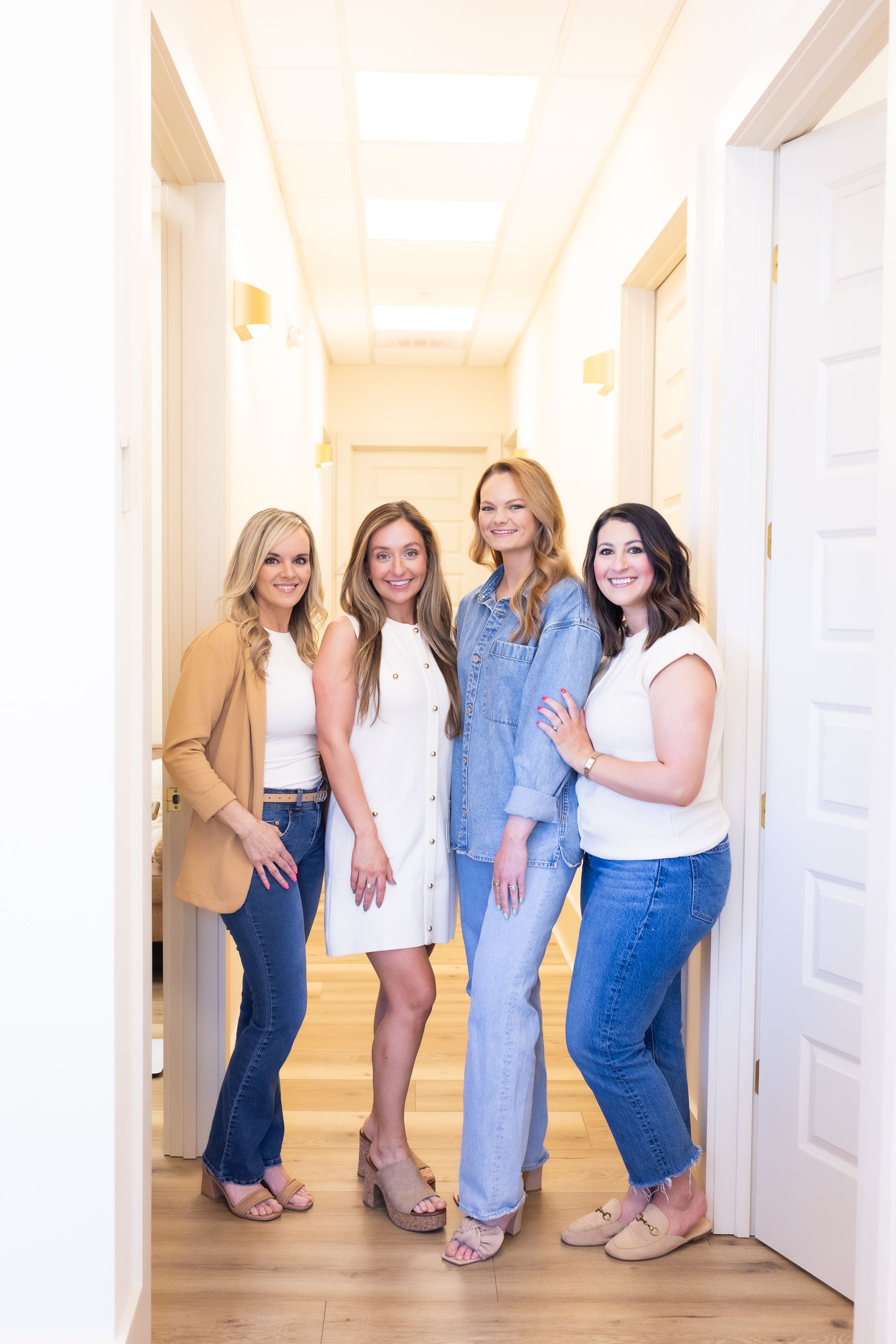 Four women stand in a bright hallway. They are dressed in casual outfits, smiling at the camera.