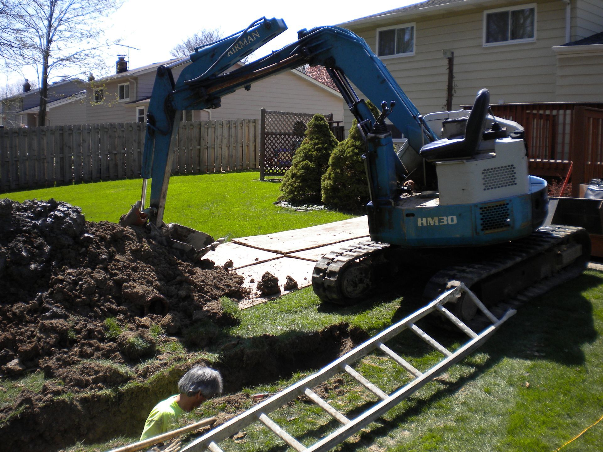 Blue excavator digging a trench in a grassy backyard. A person is working in the trench, with a ladder nearby.
