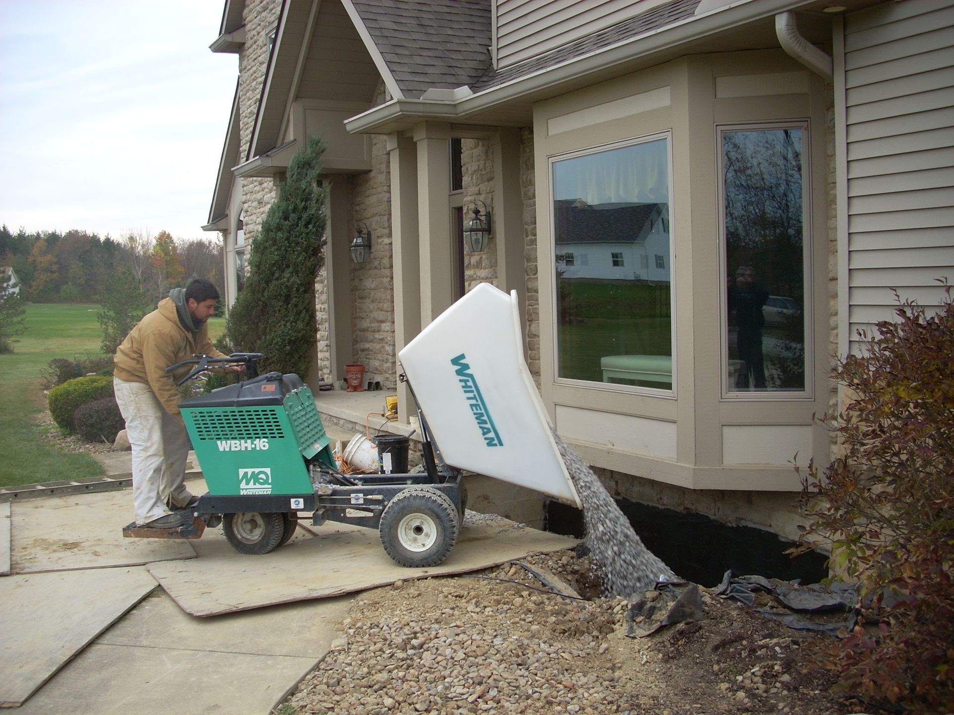 Man using a machine to remove gravel from the side of a house near a bay window.