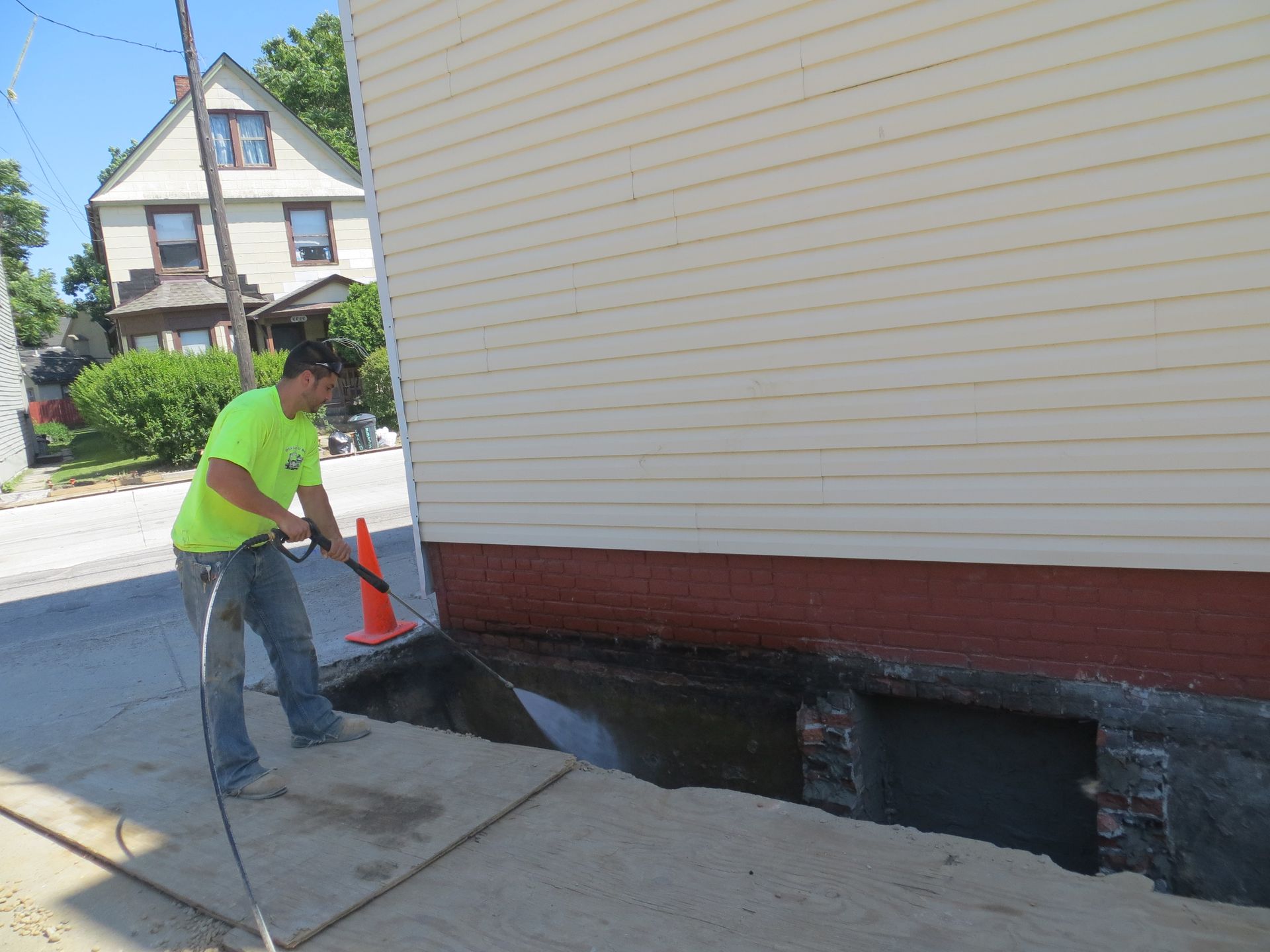 Man pressure washing a building's exterior. Beige siding, red brick foundation, sunny day.