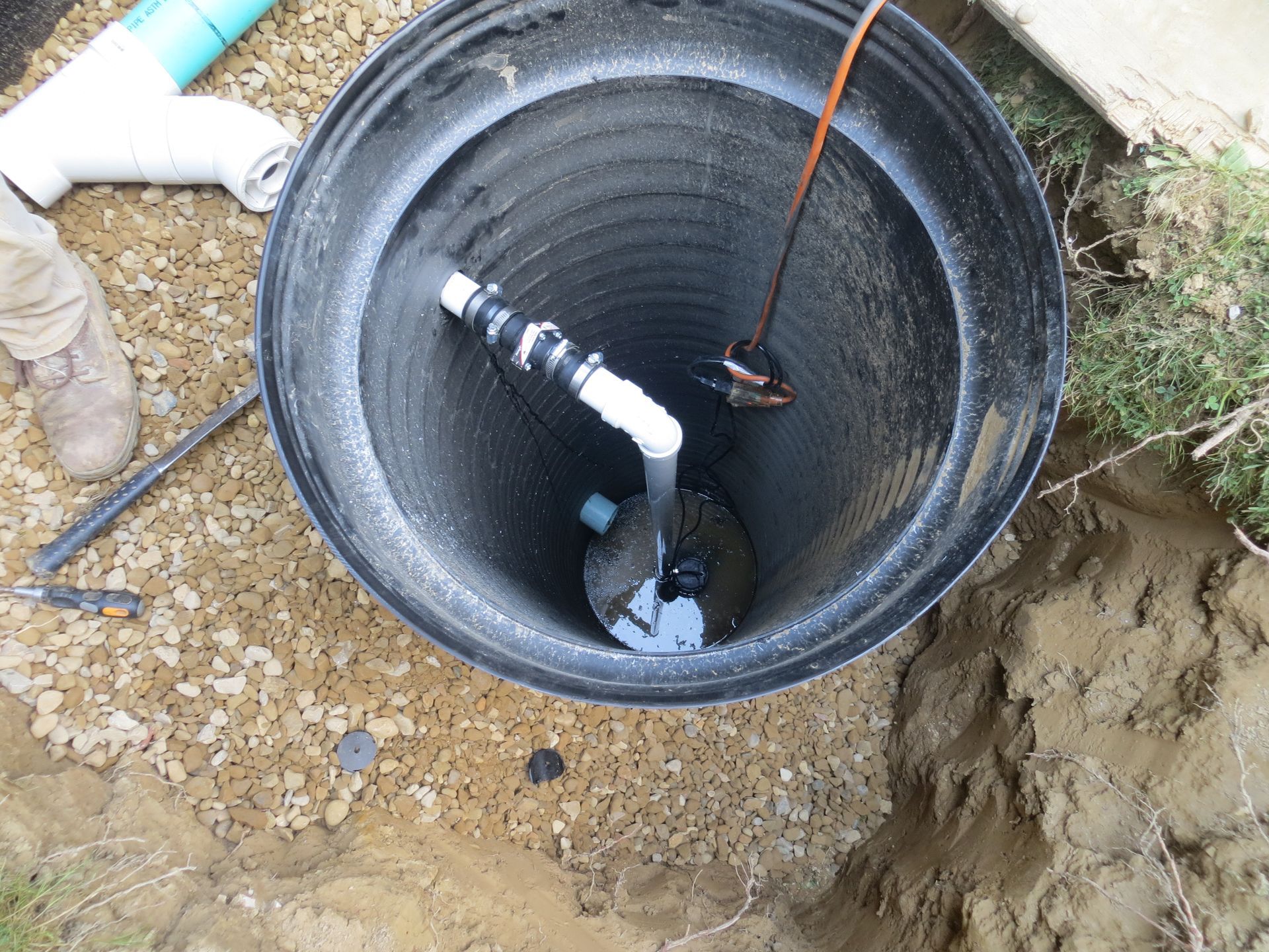 Black sump pump pit with pump and pipe installed, surrounded by gravel and dirt.