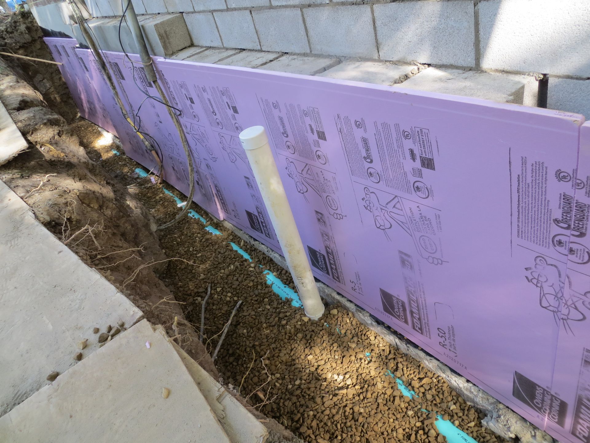 A trench next to a cinder block wall, with pink insulation, a white pipe, and gravel.