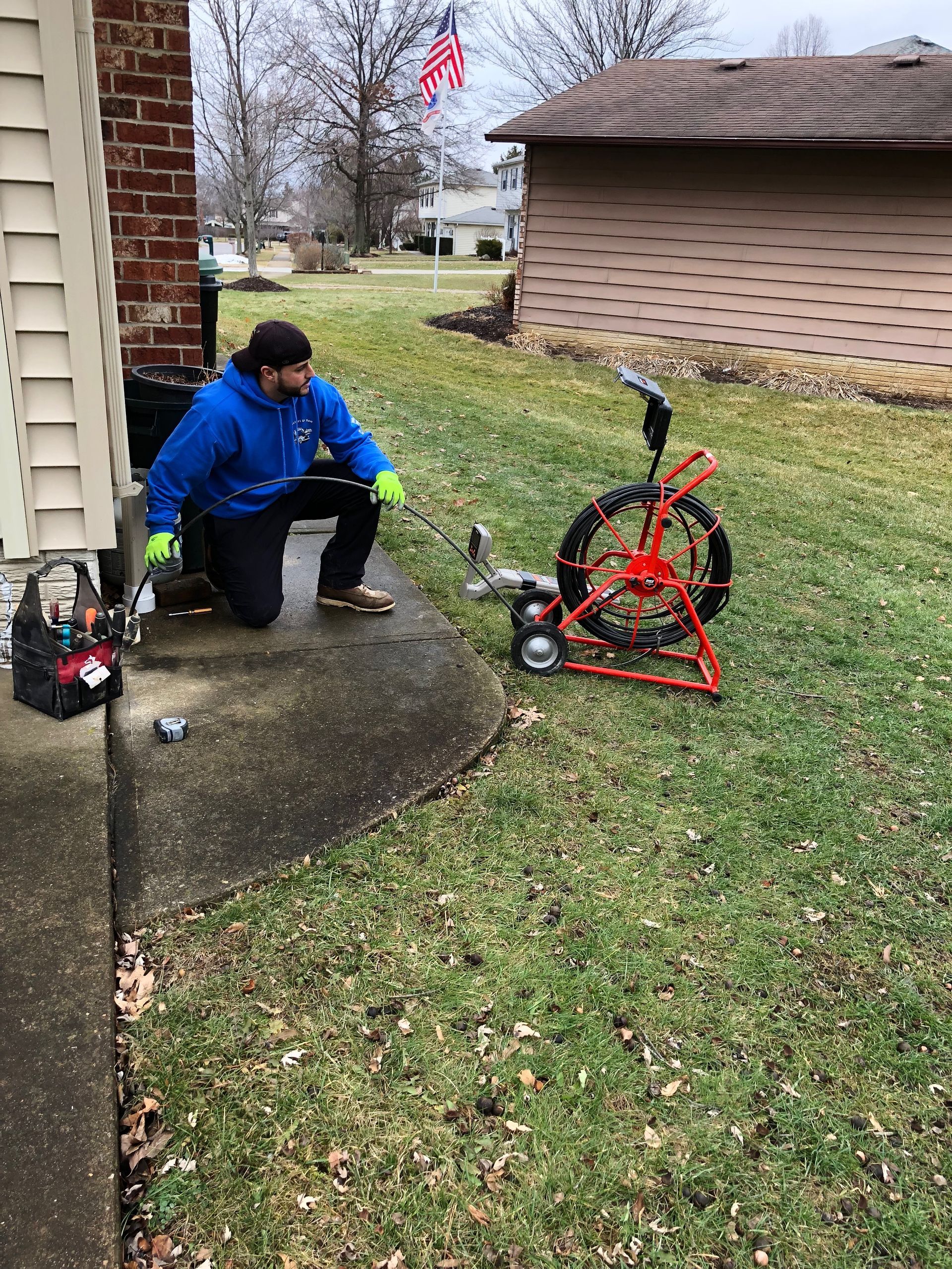 Man kneels on a porch, inspecting a drain cleaning machine, in a yard with an American flag.