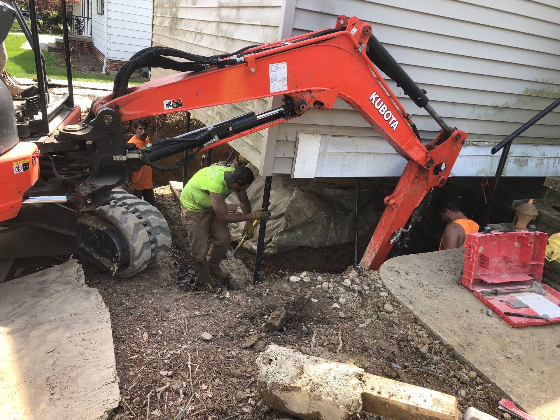 Construction crew working under a building with orange excavator. Workers in green and orange vests digging.