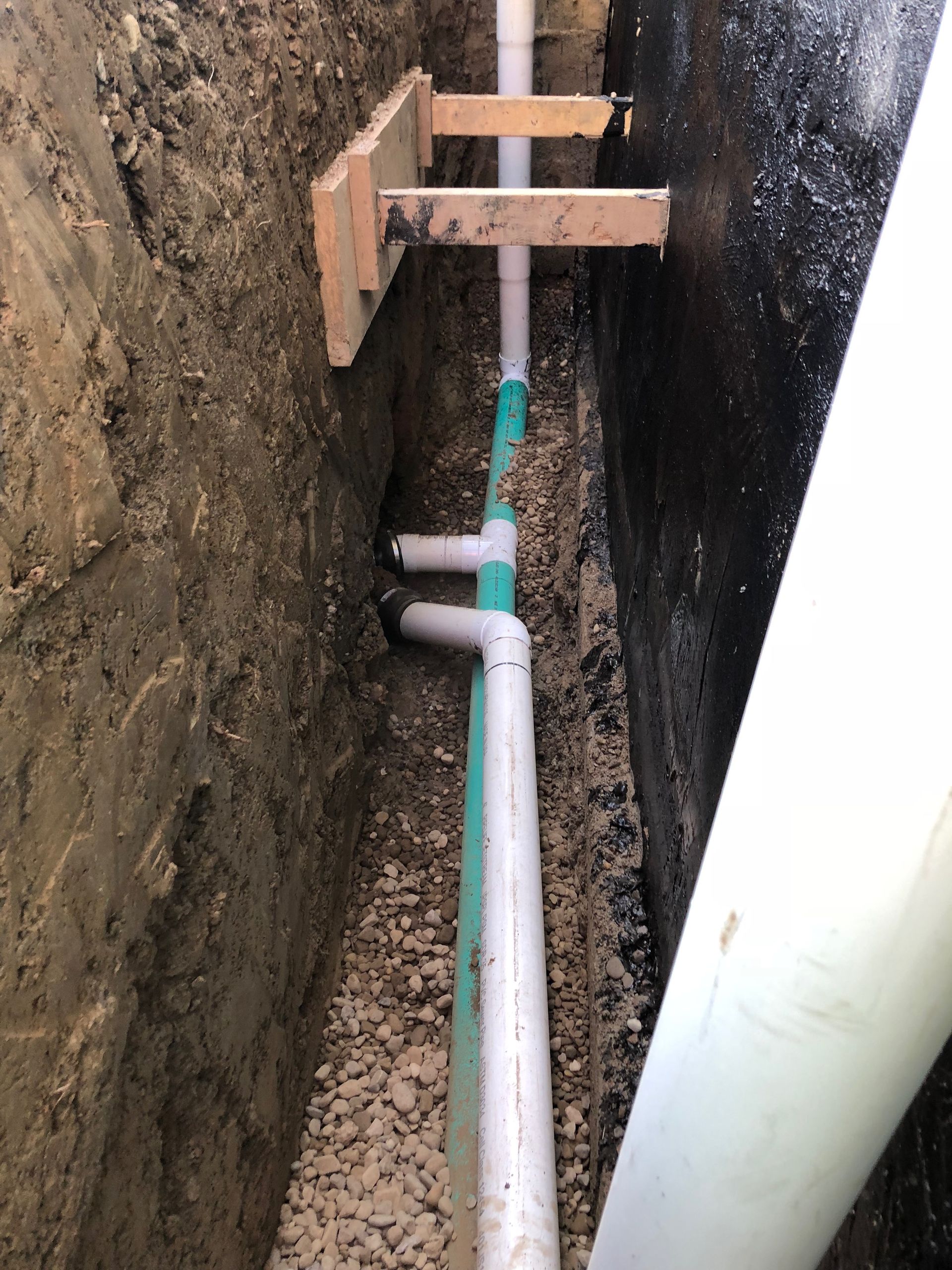 Trench with green and white PVC pipes. Wooden supports hold pipes in place. Dark, gravel-lined trench.