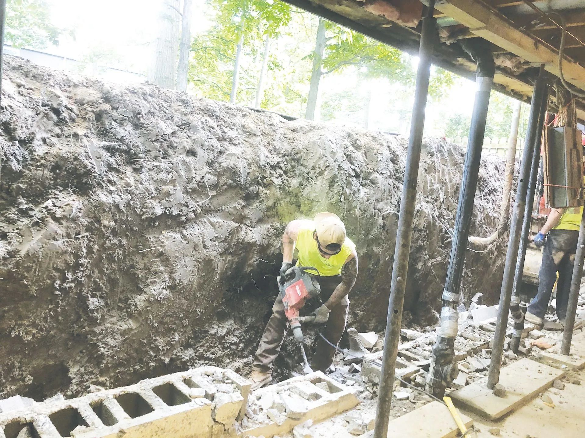 Construction worker using a jackhammer on concrete blocks, with supports and dirt wall in the background.