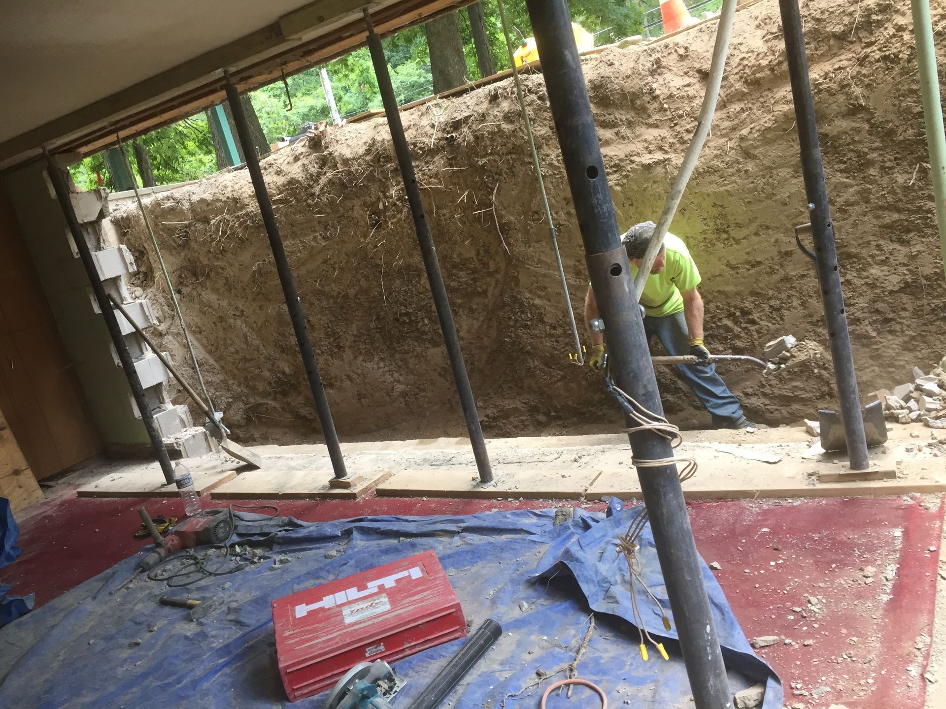 Construction worker digging in an excavated area, supported by metal posts. Red toolbox and blue tarp visible.