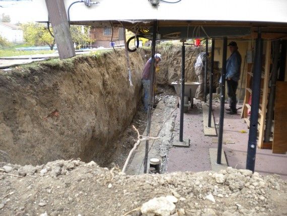 Construction workers excavating a trench next to a building, supported by metal beams.
