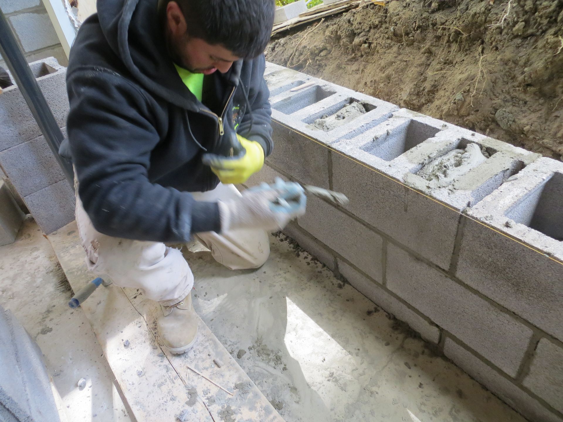 A construction worker in a black jacket is laying cinder blocks, using a trowel to apply mortar.