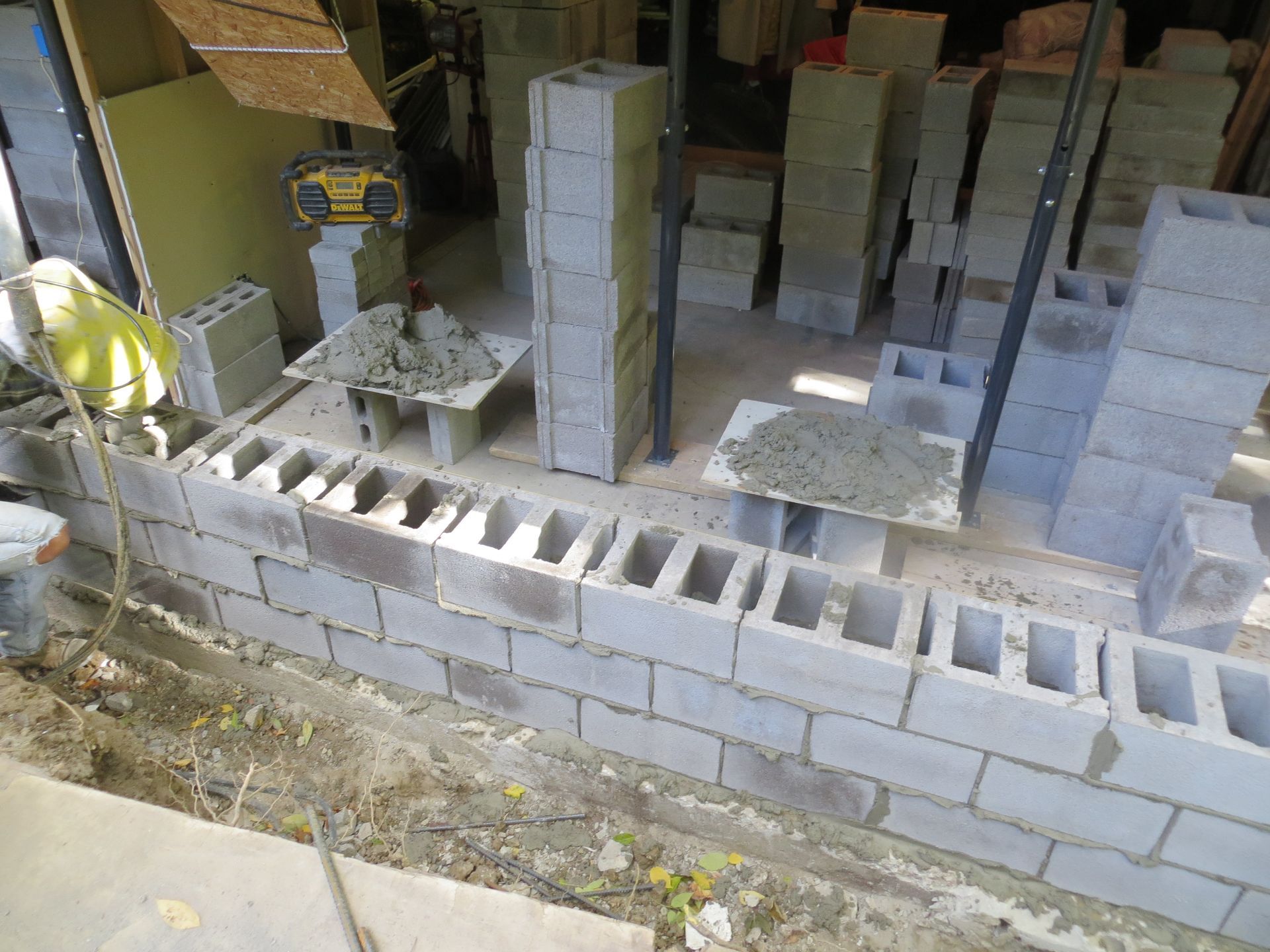 Concrete block wall under construction, indoors. Blocks stacked with mortar, various tools and materials nearby.