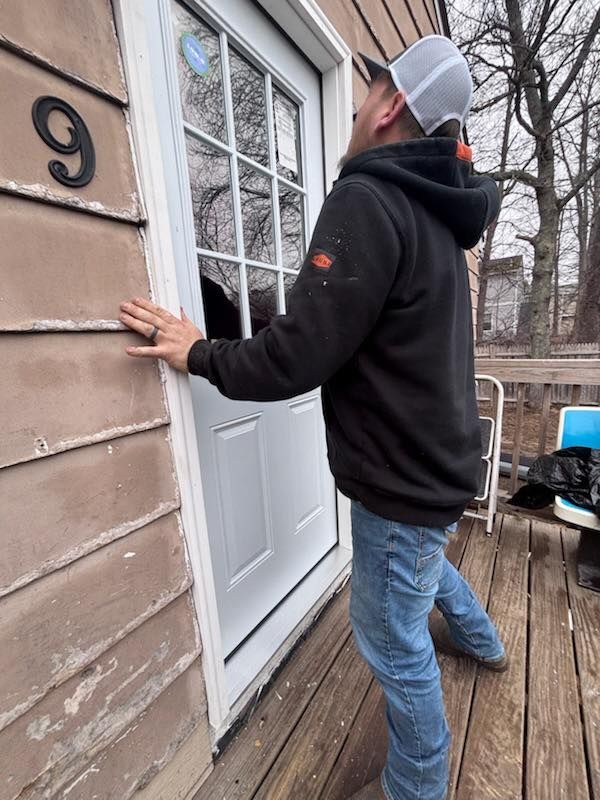 Man in jeans and a hooded sweatshirt leans against a white door with a window, on a wooden deck.