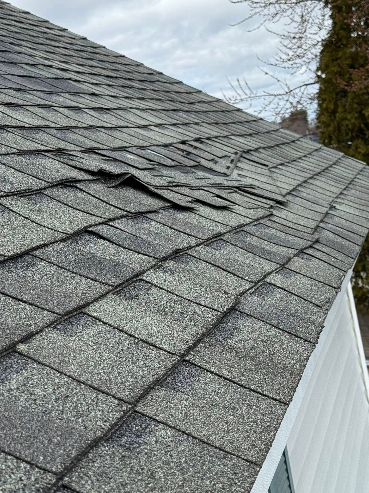 Damaged asphalt shingle roof, with missing and lifted shingles, on a white house under a cloudy sky.
