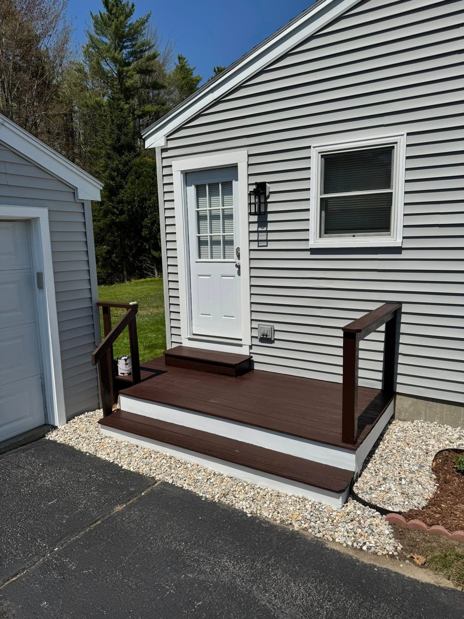 Small deck with steps leading to a white door; brown steps, white trim, grey siding, and gravel edging.