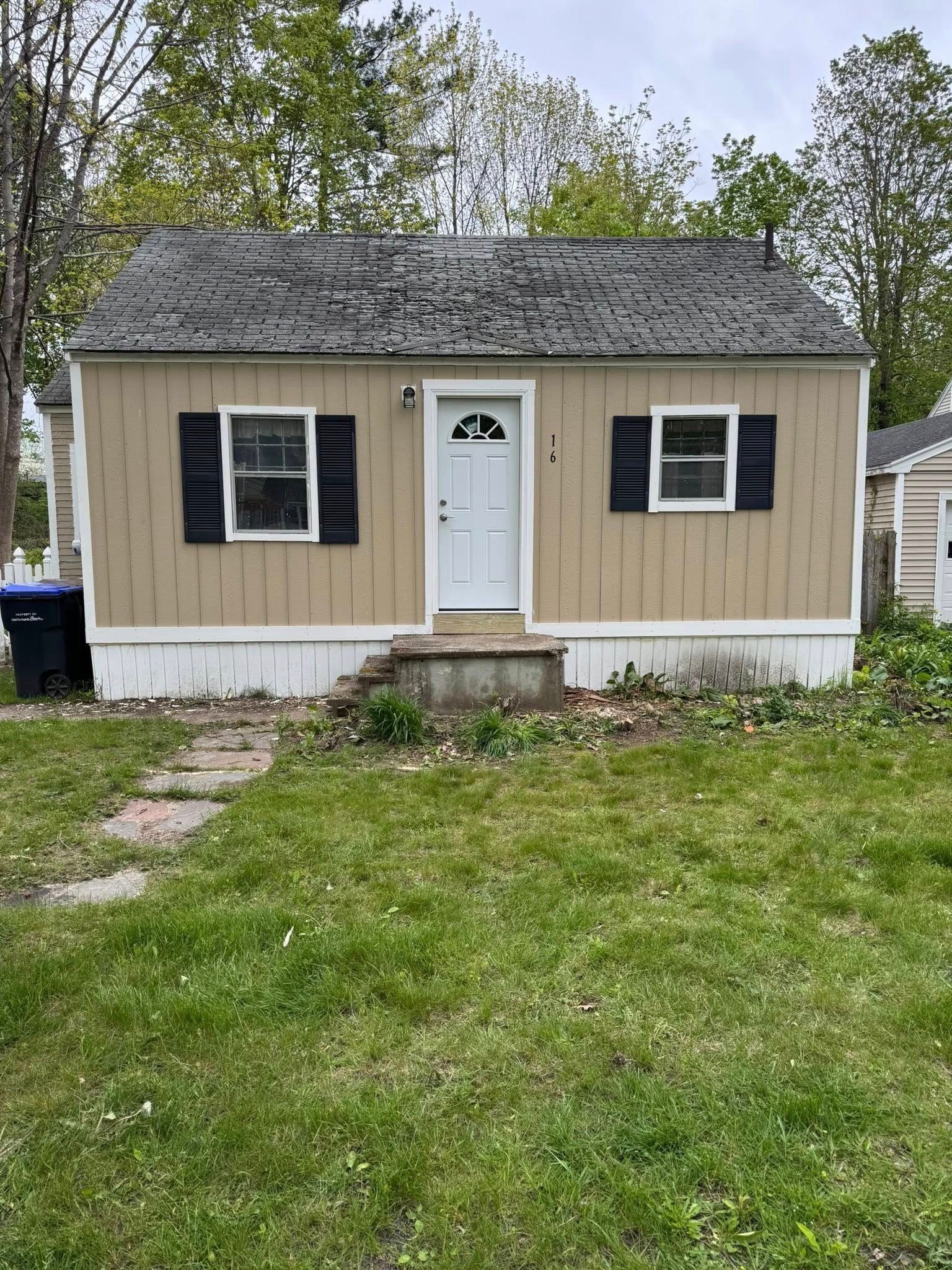 Small beige house with black shutters, white door, and overgrown yard.