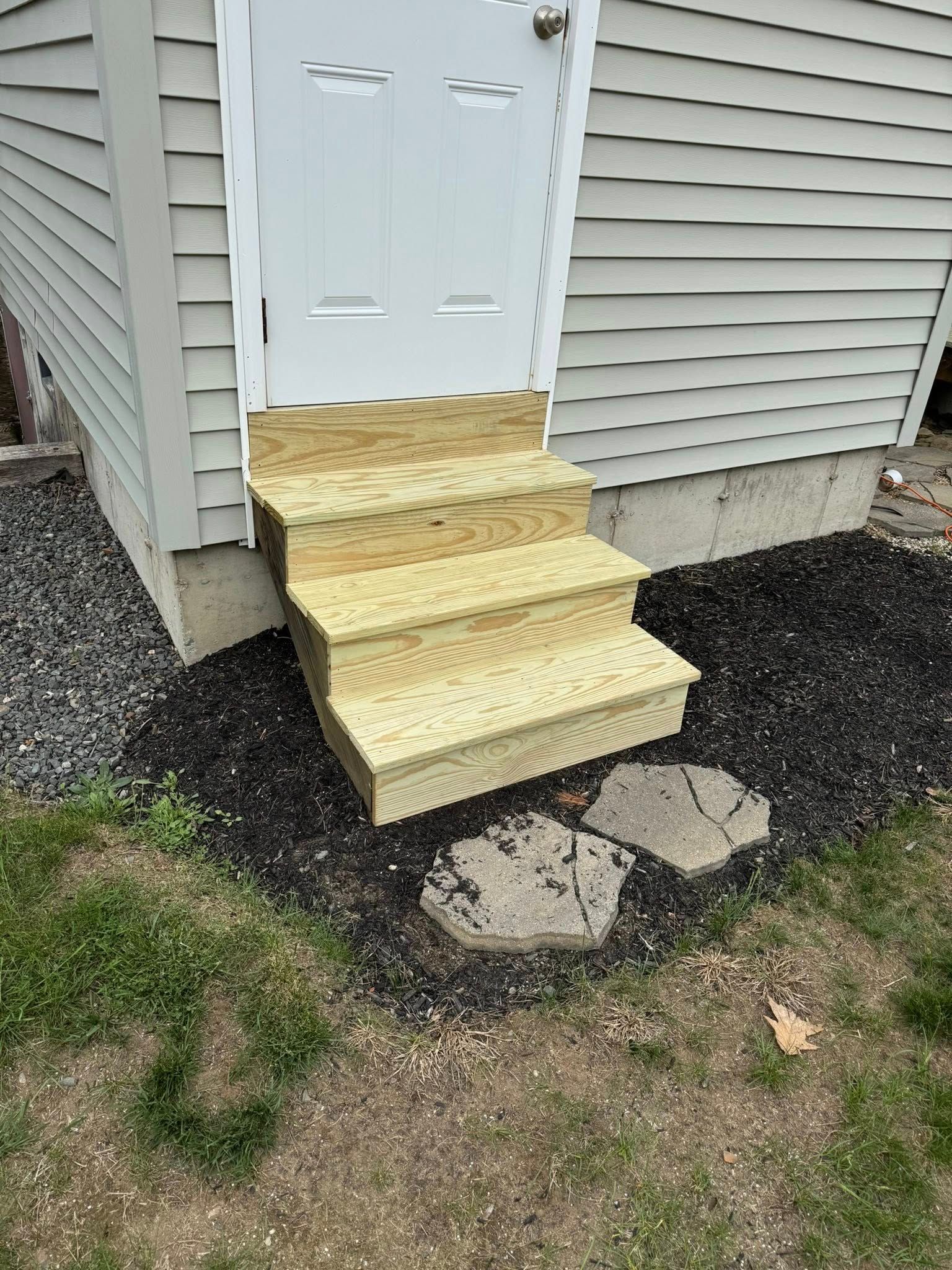Wooden steps leading up to a white door on a building with gray siding, surrounded by mulch and grass.