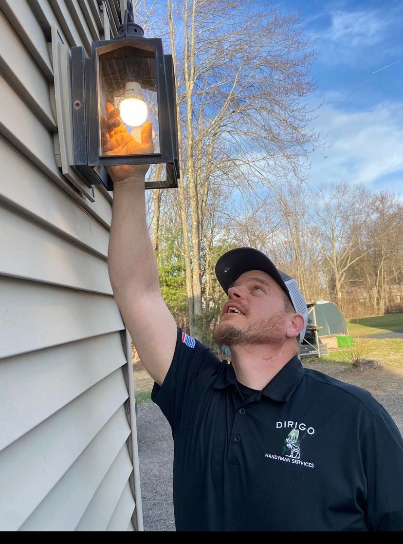 Man checking a light fixture attached to a house. He wears a hat and polo shirt. Outdoors.