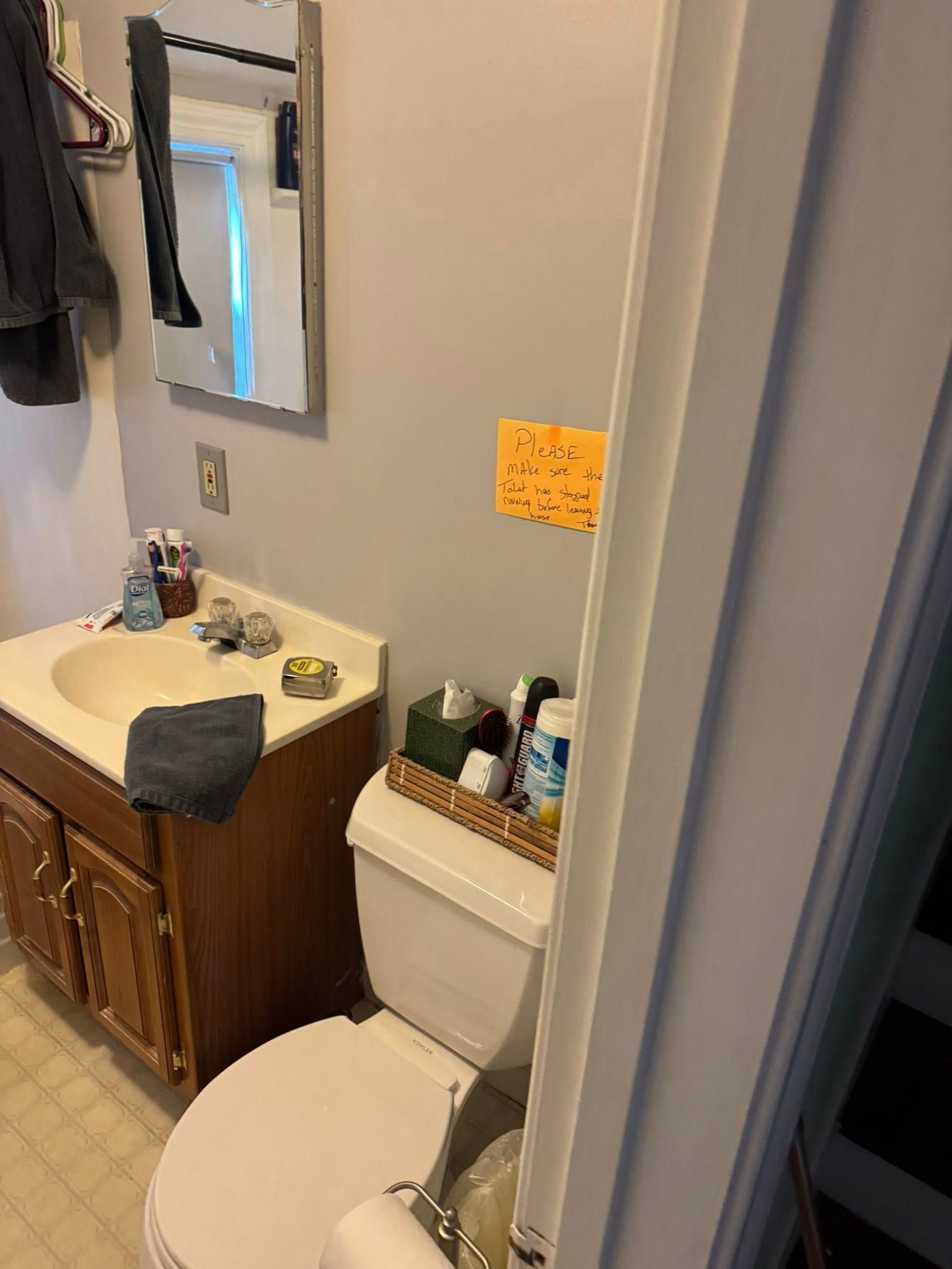 Bathroom with toilet, sink, mirror, and toiletries. Light gray walls, dark wood cabinets, and tan floor tiles.