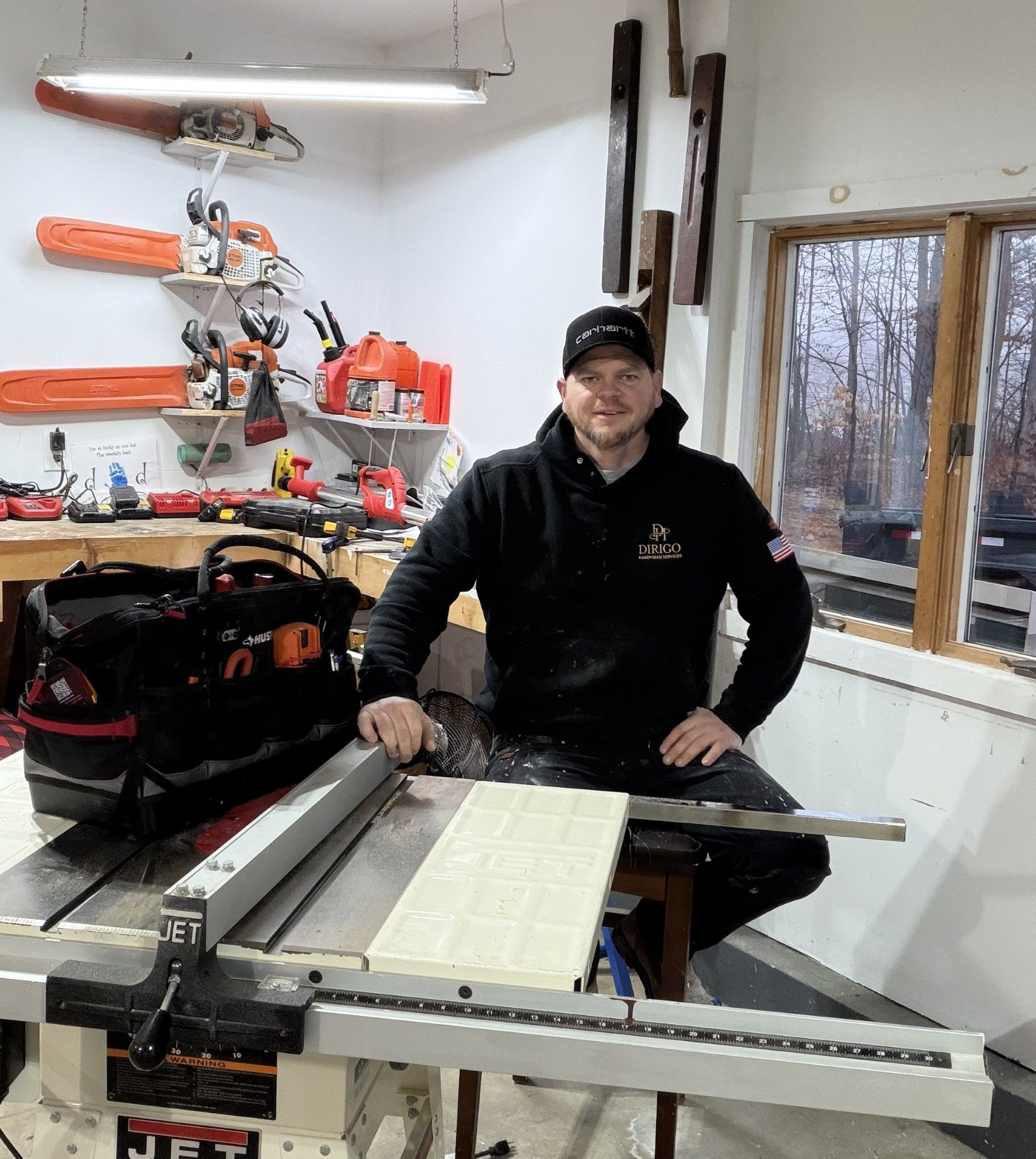 Man in workshop with a table saw, wearing black cap and sweatshirt, working with wood.