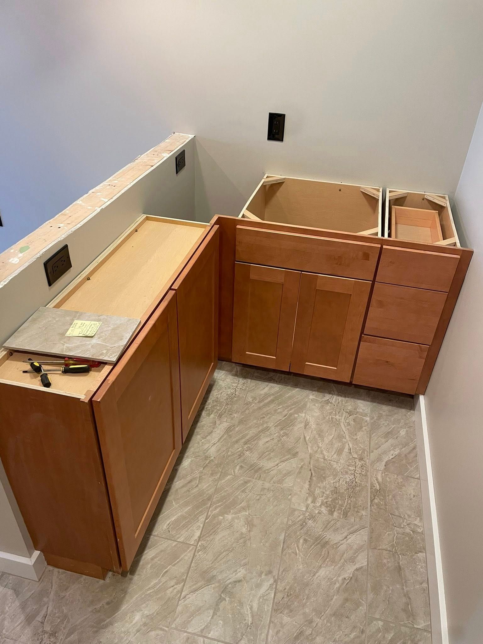 Kitchen cabinets being installed in the corner of a room, unfinished. Light brown wood, tile floor, white walls.
