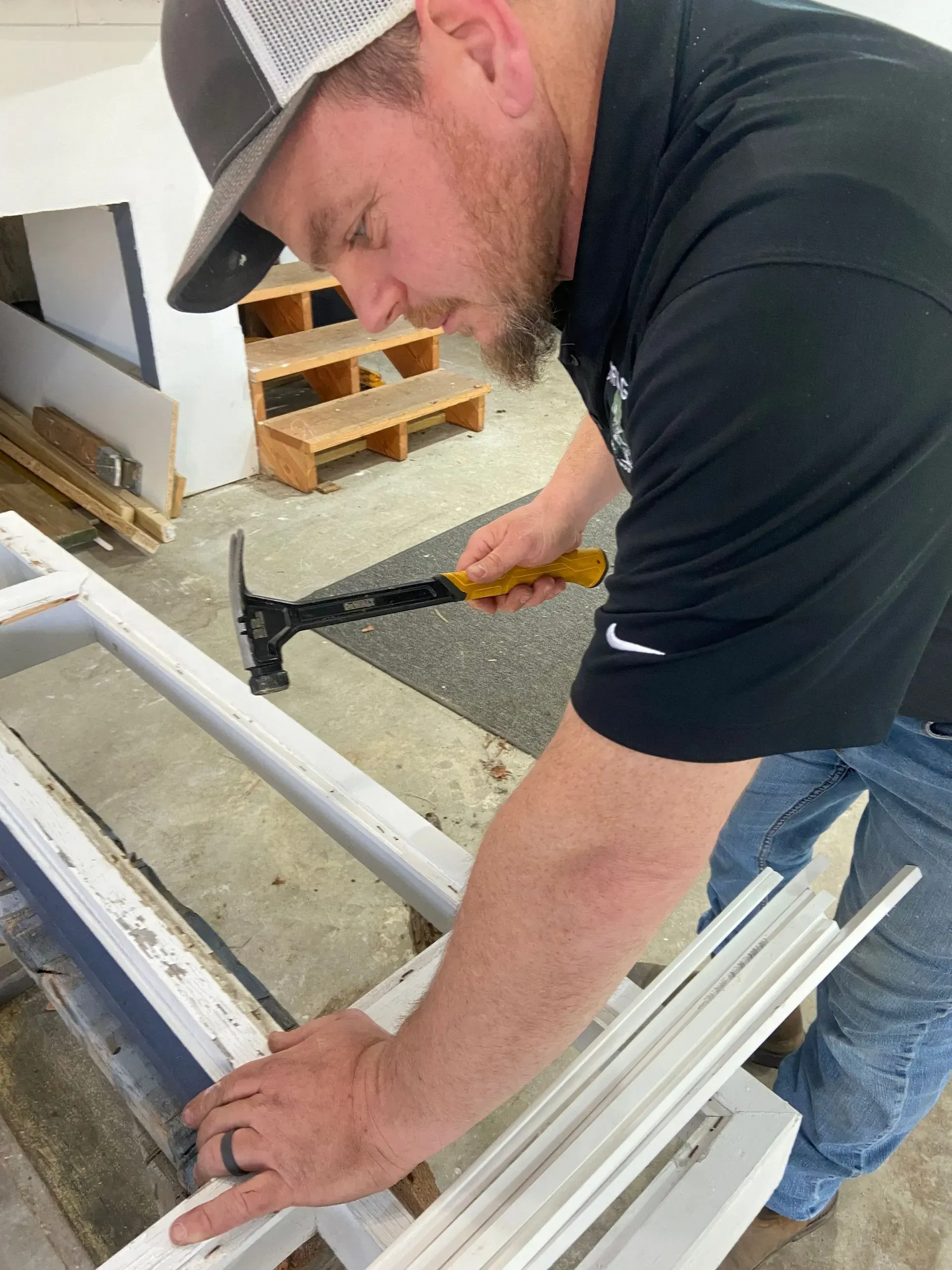 A person hammering wood trim in a workshop. Wearing a hat and black shirt, using a yellow and black hammer.