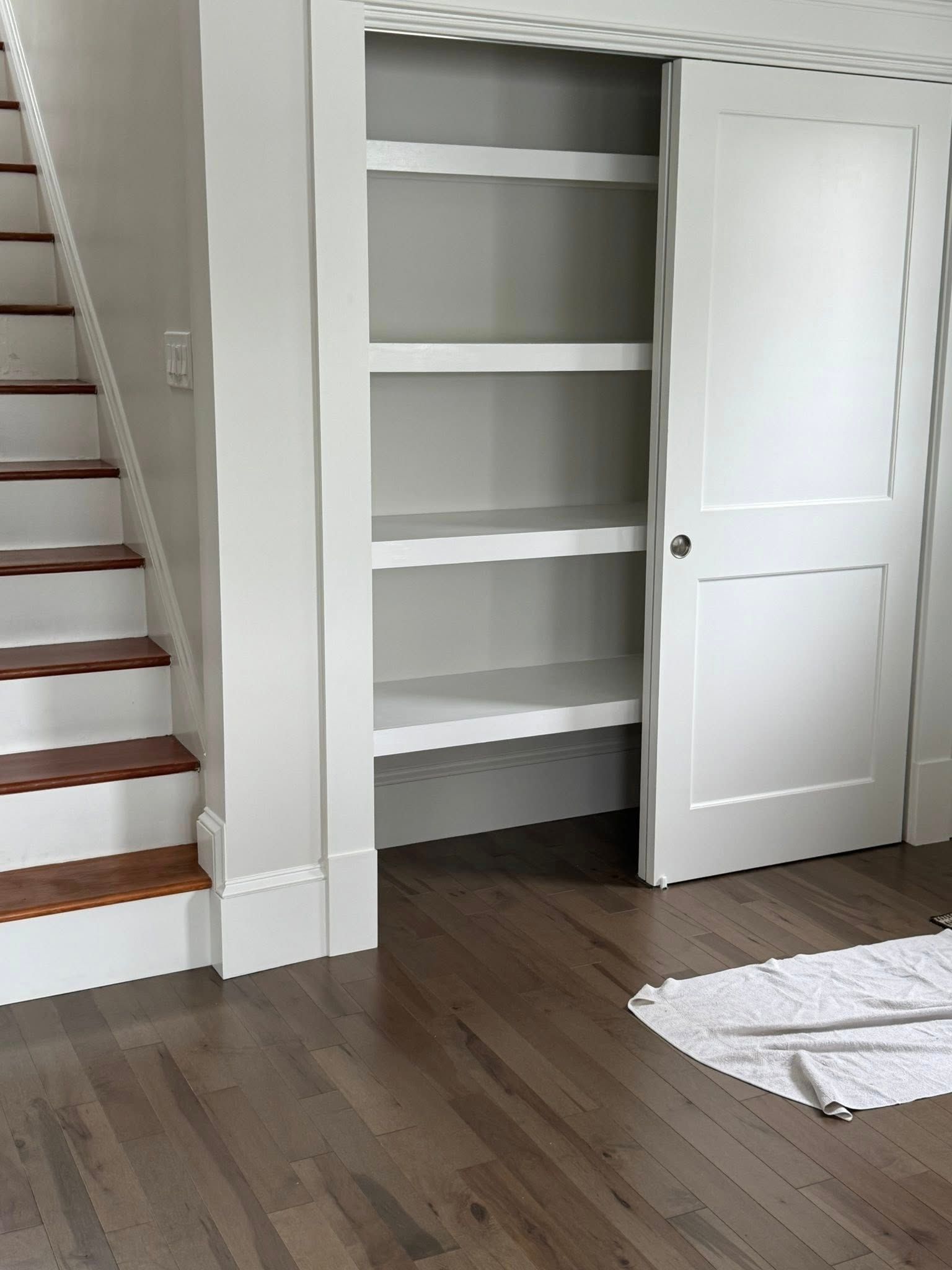 Built-in white shelves in a hallway, next to a staircase. A white door is partially open. Hardwood floors.