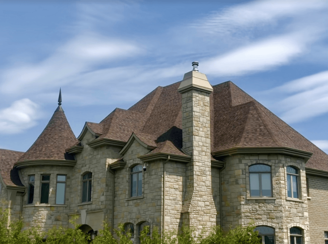 Manoir en pierre au toit brun, avec tourelle et cheminée, se détachant sur un ciel bleu nuageux.
