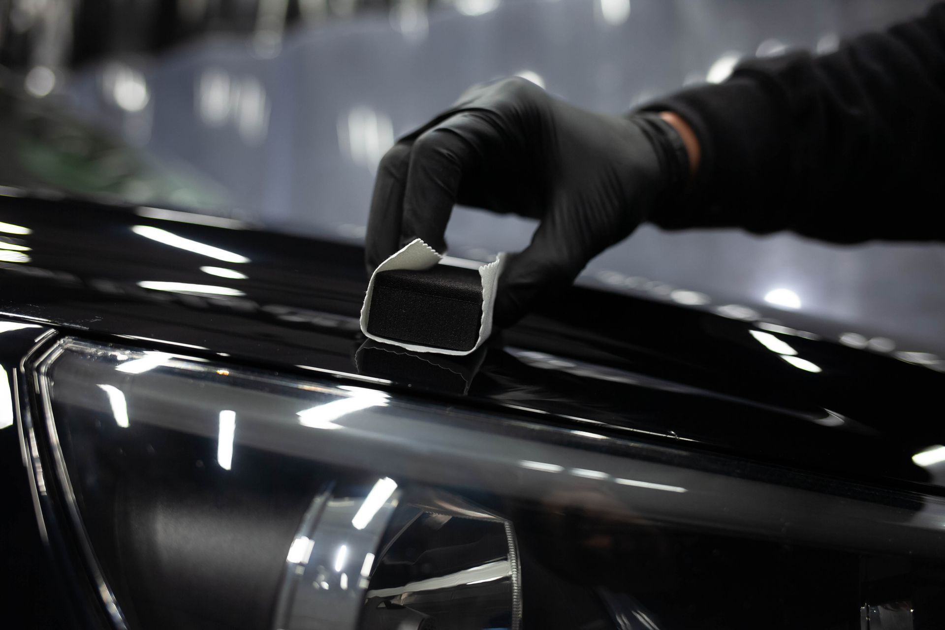 A person wearing black gloves is polishing a black car with a sponge.