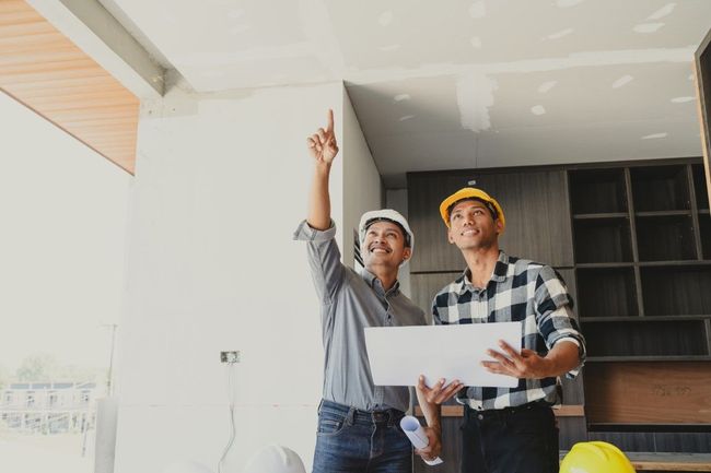 Two construction workers in hard hats examine a ceiling; one points upward, holding plans.