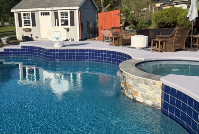 A dark-bottomed swimming pool with blue water surrounded by a grey stone border and a white fence.
