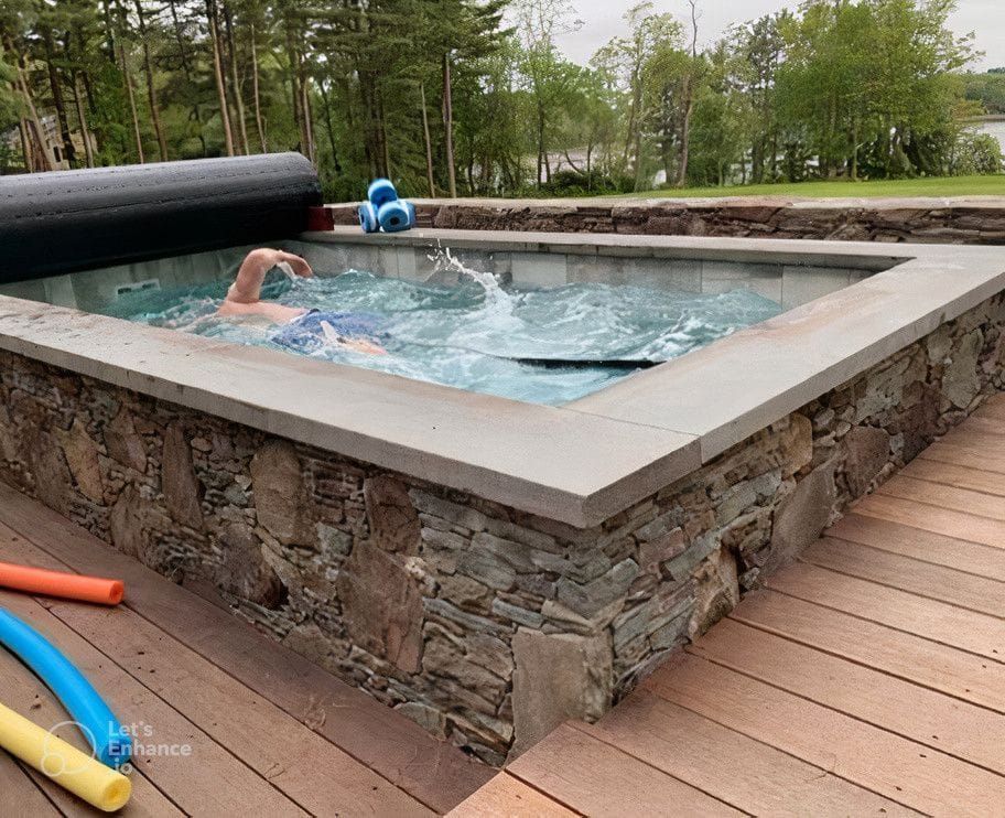 Person swimming in a small outdoor pool surrounded by stonework and wooden decking; trees in the background.