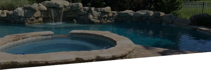 Pool with waterfall and connected spa. Rocky edges, dark teal water, green grass background.