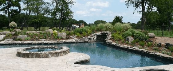 A pool and hot tub with stone accents surrounded by lush landscaping.