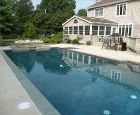 Swimming pool in a backyard with house and patio; blue water, gray patio, green trees.