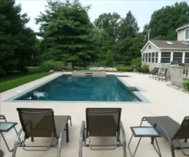 Lounge chairs face a rectangular pool. A house and trees surround the backyard pool area.