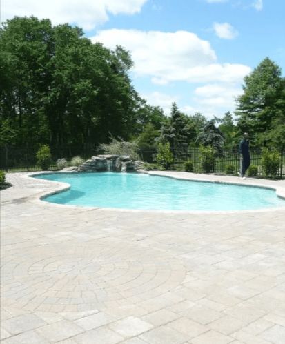 Swimming pool with waterfall, surrounded by stone patio and greenery under a blue sky.