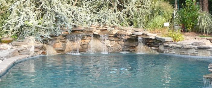 Pool with waterfalls flowing from a rock wall into the blue water. Lush green plants surround the area.